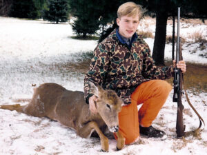 teenage boy kneeling in a snow-covered field with one hand on a rifle and the other on a dead whitetail deer